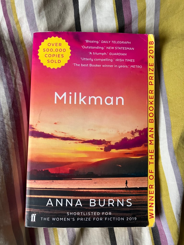 A copy of the book Milkman by Anna Burns, lying on some striped material. the cover shows a sunset above a lake, a lone female figure walking on the shore, hills in the distance. There is a yellow splash detail saying; OVER 500,00 COPIES SOLD