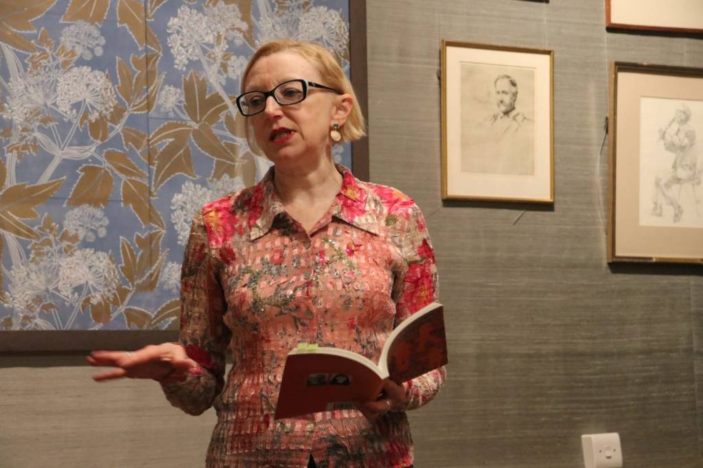 Hilaire standing in front of a grey wall with several art works behind her. She is wearing a crinkly pink shirt with an abstract floral pattern on it. In her left had she's holding open a book and her right hand is gesturing as she reads.