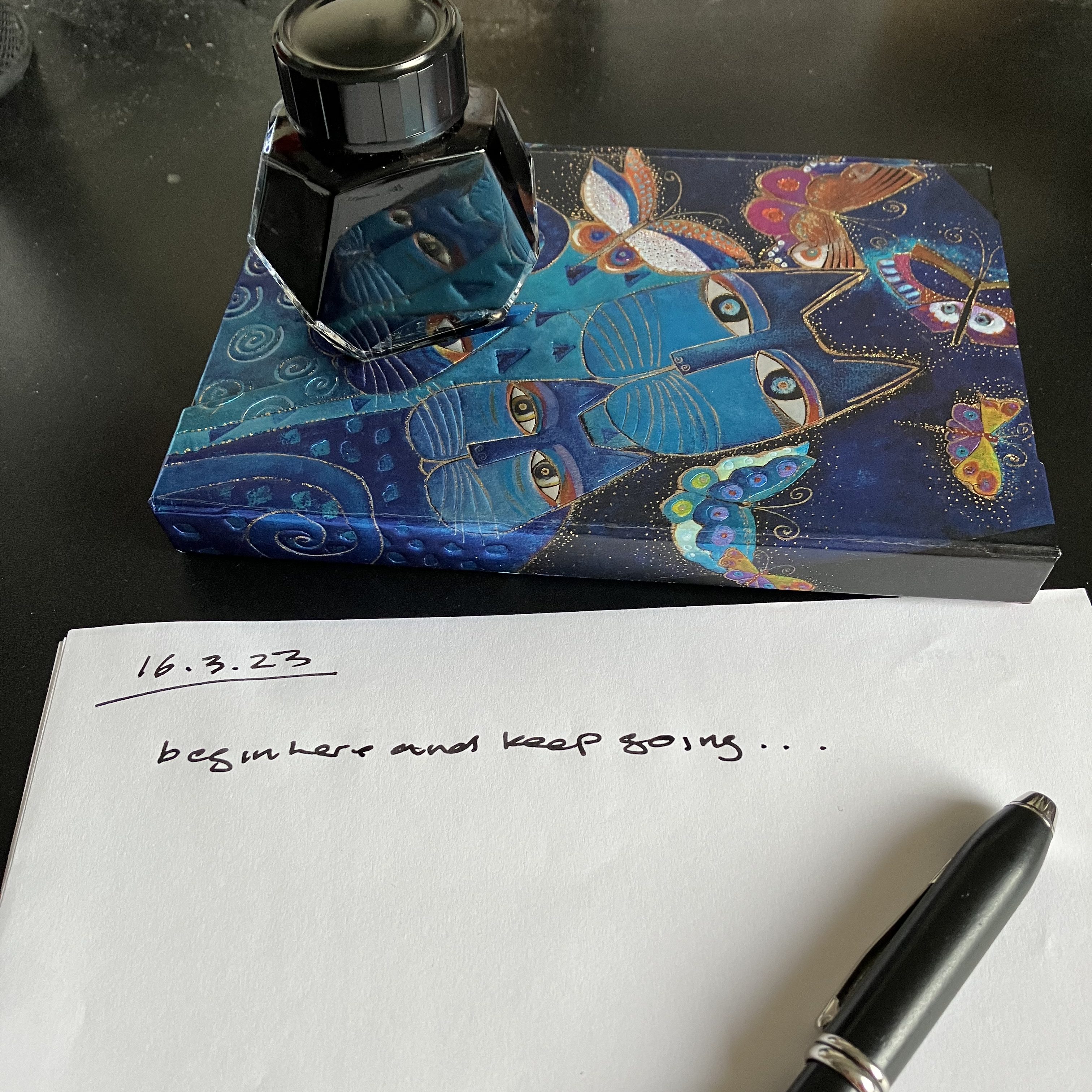 A plain piece of paper, the date hand-written in black ink on the top lefthand corner: 16..3.23. The date is underlined. Beneath that id=s hand written: begin here and keep going ...
Above the page on a black desk is a hardback notebook with a think dark blue cover and Art Deco style cat images. A black bottle of ink sits on top of the notebook.