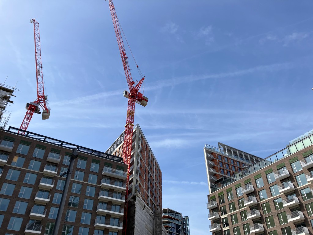 A view if the tops of several tall, angular apartment blocks, and two red construction crane, with blue sky and streaks of white cloud behind.