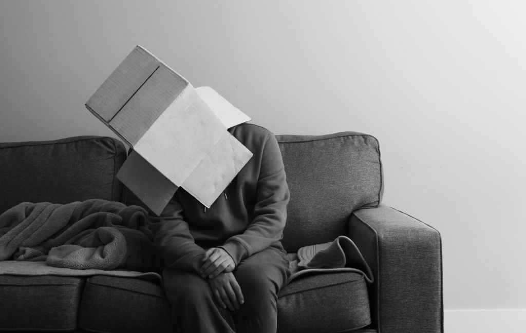 A black and white photo of a man sitting on a sofa with a cardboard box on his head.