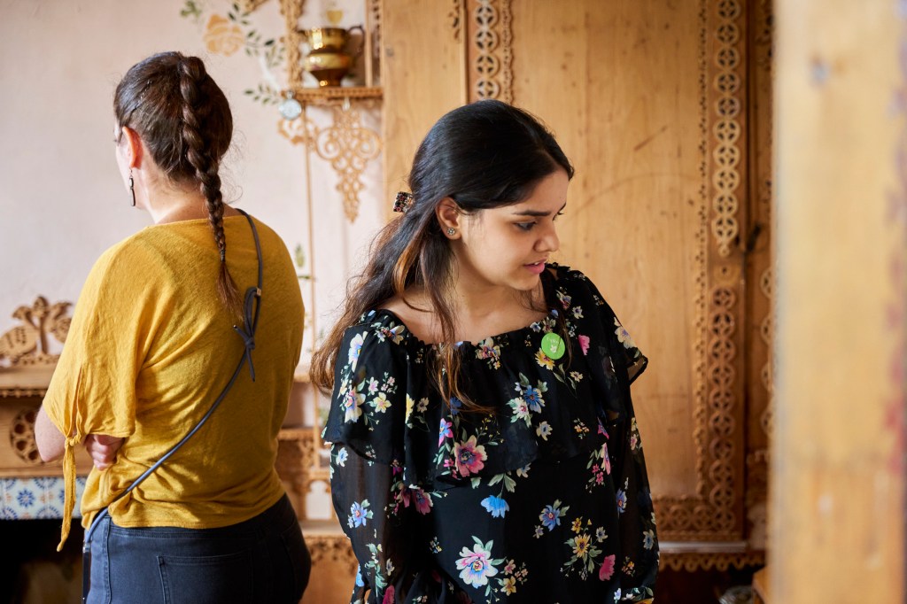 Two women standing in a room, one with her back to the camera and the other looking slightly downwards to the right. Behind them there is carved fretwork on a wooden wardrobe, stencilled flower and leaf patterns on a cream wall, and more carved fretwork above a fireplace. A lustreware jug stands on a small plinth attached to the side of the wardrobe.