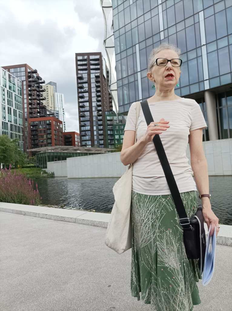 Hilaire stands in front of the U.S.Embassy & its moat. Tall buildings are visible in the middle distance on the left hand side. Hilaire is speaking & gesturing with her left hand.