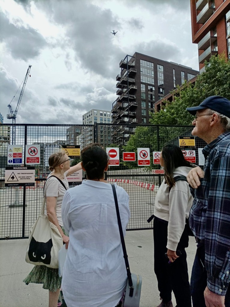 Hilaire and three others are seen from behind, standing in front of a site entrance gate with safety signs on it. On the right hand side tops of a few trees are visible as well as tall apartment blocks. The sky is cloudy, and a plane passes high overhead. A tall crane is visible in the middle distance.