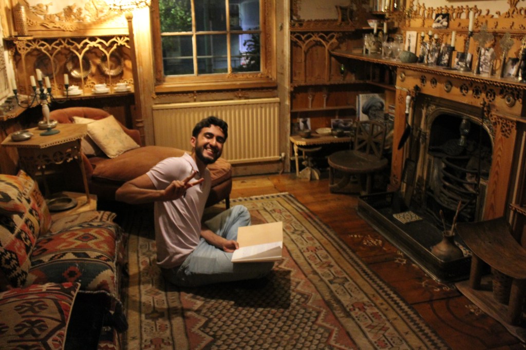 A young man sits cross-legged on a kilim rug with an open exercise book in his lap. He is smiling and making a peace sign with his rihgt hand towards th camera. In the wall opposite him is a disused fireplace and on the mantelpiece above are many candlesticks and other objects