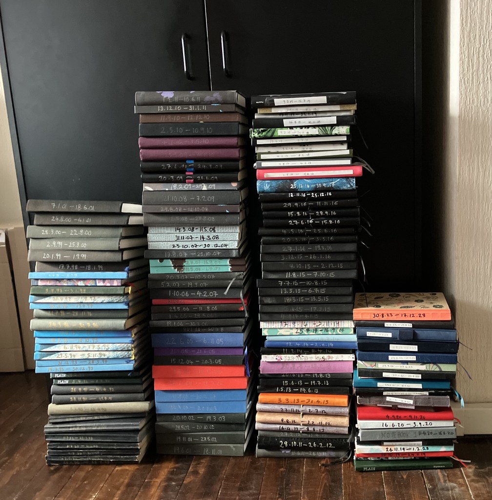 Four stacks, of varying height, of mostly A5 size journals, standing in front of a black wardrobe. The journals are stacked horizontally, spines outwards, with dates written on them in black against white stickers or gold or silver pen directly on the spine. The covers are mostly black, but some are blue, purple, red or multicoloured
