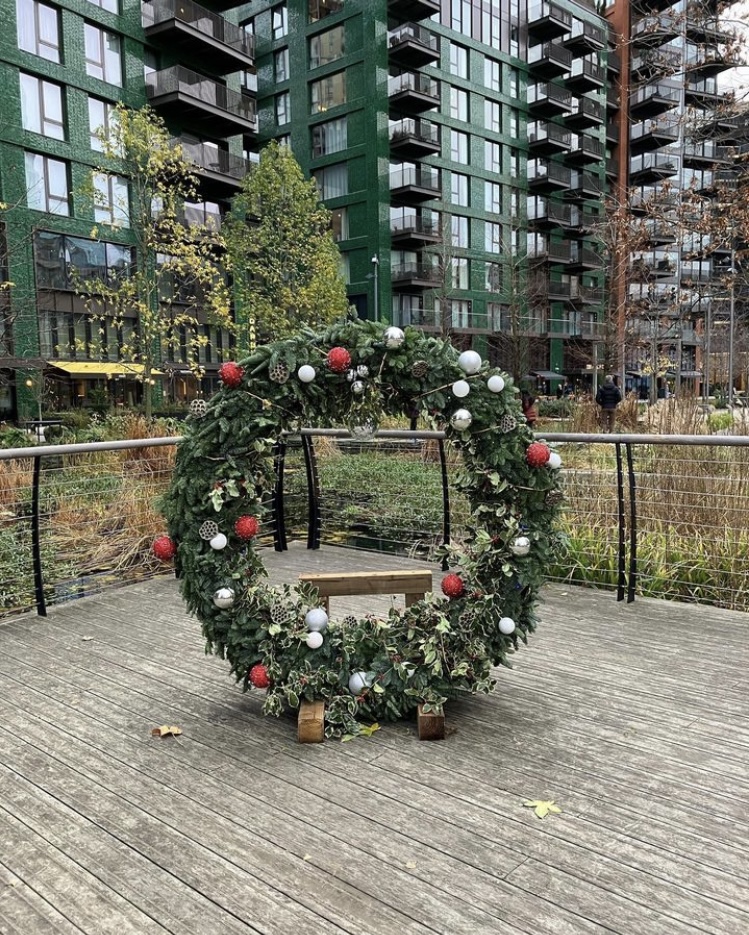A large Christmas wreath stands in some wooden decking. Beyond the decking is a bed of reds and in the background tall apartment blocks