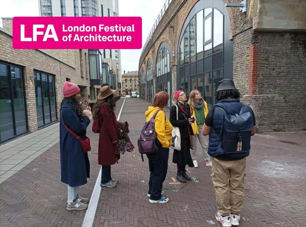 A group of people standing on a brick path looking up at a Victorian railway viaduct. Behind them is a low-rise brick and glass building and beyond that a tall new build residential building.