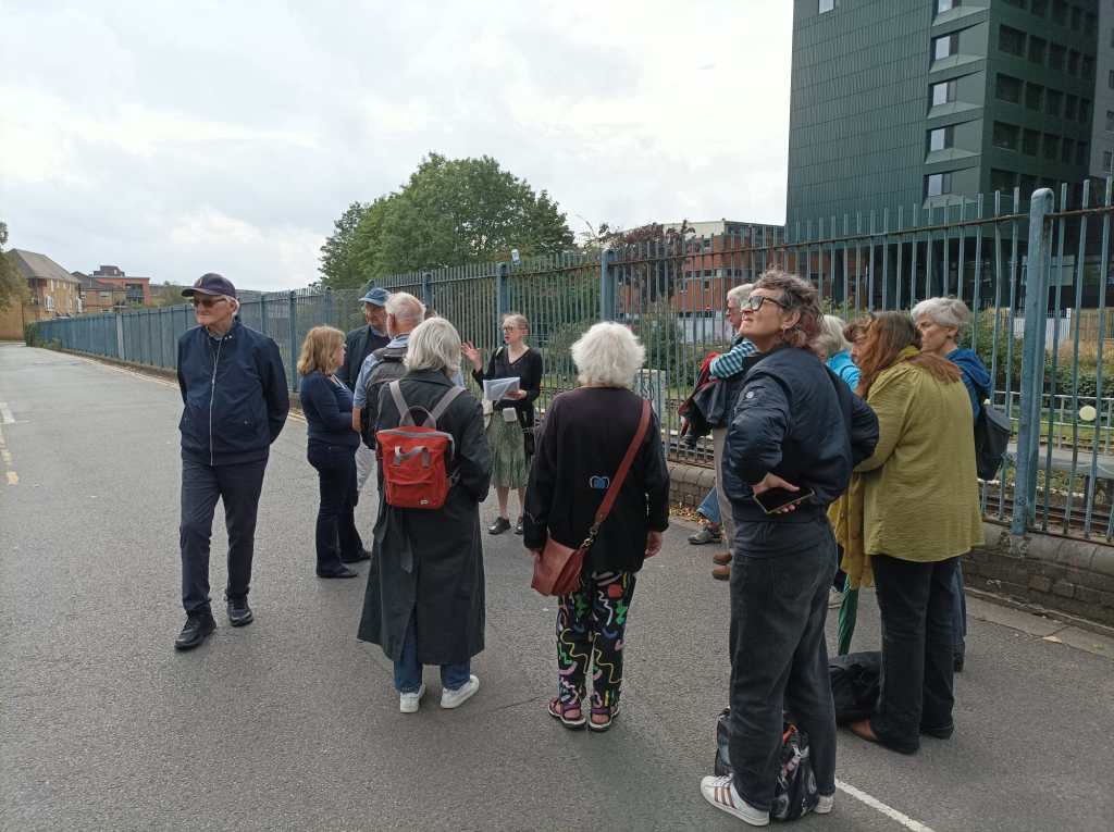 A group of people standing looking to the left as Hilaire speaks. Behind them on the right is a railway line and a new tall building