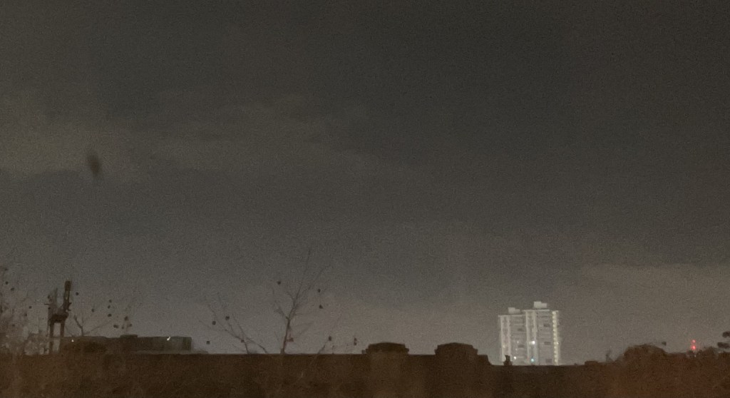 a dark night sky with a tower block in the distance and the tops of bare trees in the foreground